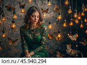 Portrait of a beautiful girl in a green dress among many suspended light bulbs and butterflies fluttering around her. Daydreaming. A magical atmosphere. Стоковое AI-изображение, AI-художник Photojuli / Фотобанк Лори