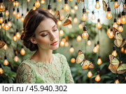 Portrait of a beautiful girl in a green dress among many suspended light bulbs and butterflies fluttering around her. Daydreaming. A magical atmosphere. Стоковое AI-изображение, AI-художник Photojuli / Фотобанк Лори