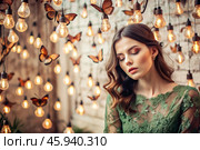 Portrait of a beautiful girl in a green dress among many suspended light bulbs and butterflies fluttering around her. Daydreaming. A magical atmosphere. Стоковое AI-изображение, AI-художник Photojuli / Фотобанк Лори
