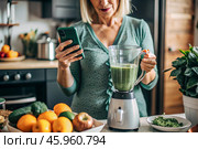 An elderly woman in the kitchen is looking at healthy food recipes on her phone and preparing a vegetable smoothie made from broccoli in a blender. Healthy detox diet drink. Стоковое AI-изображение, AI-художник Photojuli / Фотобанк Лори