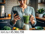 An elderly woman in the kitchen is looking at healthy food recipes on her phone and preparing a vegetable smoothie made from broccoli in a blender. Healthy detox diet drink. Стоковое AI-изображение, AI-художник Photojuli / Фотобанк Лори