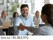 Young businessman shakes hands with a colleague during a networking event in a bright office, smiling confidently amidst engaged attendees. Стоковое AI-изображение, AI-художник Sergey Grishin / Фотобанк Лори