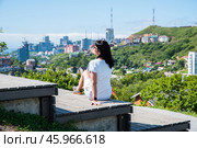 A 50-year-old Russian woman in a white dress sits on wooden steps in Nagorny Park, Vladivostok, enjoying the cityscape and greenery under the summer sun. Стоковое фото, фотограф Татьяна Терехина / Фотобанк Лори