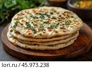 A stack of fragrant flatbreads garnished with fresh herbs. The flatbreads look soft and golden brown, with perfectly toasted crusts. They lie on a wooden round board, creating a cozy and appetizing impression. The background is slightly blurred, showing fresh herbs and sauce in the bowl, which emphasizes the culinary theme. Стоковое AI-изображение, AI-художник Анна Кривицкая / Фотобанк Лори