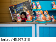 Россия, Москва, Площадь Революции - Январь 2025. Девочка за прилавком с кошками - копилками на рынке. Декорации из фильма Леонида Гайдая "Операция "Ы" и другие приключения Шурика. Фотозона. Russia, Moscow, Revolution Square - January 2025. The girl behind the counter with the piggy bank cats at the market. Scenery from Leonid Gaidai's film "Operation Y" and other adventures of Shurik. The photo zone. Стоковое фото, фотограф Мария / Фотобанк Лори