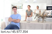 Upset young guy sitting at kitchen table on background of dissatisfied wife standing with mother-in-law behind. Family conflicts and communication problems concept. Стоковое видео, видеограф Яков Филимонов / Фотобанк Лори