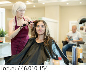 Elderly female hairstylist using curling wands to add volume for hair of young girl. Стоковое фото, фотограф Яков Филимонов / Фотобанк Лори