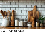 Kitchen corner with wooden and ceramic accessories. Jars with ingredients such as sugar, salt, spices and cereals, all in glass containers with lids, are placed against the background of white hog-style tiles. Nearby are wooden spice grinders, wooden cutting boards, a ceramic jar with wooden spoons and a small pot of herbs. The image conveys the atmosphere of coziness and functionality in the kitchen. Стоковое AI-изображение, AI-художник Анна Кривицкая / Фотобанк Лори