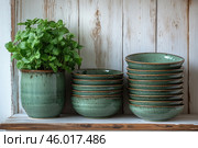 Ceramic items in rustic style. On the left is a green pot with thick fresh greenery, and next to it are neatly stacked bowls with green glaze. All the items are made in a harmonious natural style, with texture and shades that create a sense of handcrafted work. Against the background of an old wooden cabinet or a wall with a whitewashed surface, the composition looks cozy and aesthetically pleasing. Стоковое AI-изображение, AI-художник Анна Кривицкая / Фотобанк Лори