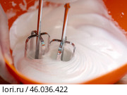 Metal mixer whisks beating a translucent egg white into a stiff white foam, set against a vibrant orange bowl, showcasing the process of baking and food preparation. Стоковое фото, фотограф Татьяна Степанищева / Фотобанк Лори