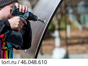 A man holds a battery-powered screwdriver and assembles a new greenhouse made of polyethylene, polycarbonate. Macro photography. Side view. Preparing for the spring garden season. Стоковое фото, фотограф Photojuli / Фотобанк Лори