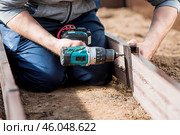A man holds a battery-powered screwdriver and collects new beds from WPC into a greenhouse made of polyethylene, polycarbonate. Macro photography. Side view. Preparing for the spring garden season. Стоковое фото, фотограф Photojuli / Фотобанк Лори