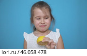 Funny little child girl try to taste fresh lemon, wrinkles and makes a grimace. Sour taste, facial emotions negative, in white dress, isolated blue background, copy space. Стоковое видео, видеограф Ирина Михайличенко / Фотобанк Лори