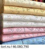 Neatly folded stacks of fabric in pastel shades: yellow, white, pink and blue. The fabric has an openwork pattern consisting of small holes and embroidery, which gives it airiness and sophistication. It resembles cotton fabric with Richelieu embroidery or lace embroidery, which is used to create clothing, bed linen and decorative interior elements. Стоковое AI-изображение, AI-художник Анна Кривицкая / Фотобанк Лори