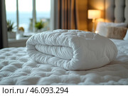 Cozy bedroom with warm lighting. In the center of the frame lies a neatly folded white blanket, laid on a bed with a soft, voluminous bedspread. The background is slightly blurred, in the background you can see a large window through which natural light penetrates. The interior includes decorative pillows, a bedside lamp and decorative elements that create an atmosphere of coziness and tranquility. Стоковое AI-изображение, AI-художник Анна Кривицкая / Фотобанк Лори
