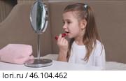Fashion little girl in a white T-shirt is sitting at the dressing table in front of the mirror and applying makeup with lipstick. Advertising of children's cosmetics concept. Footage for kids blog. Стоковое видео, видеограф Ирина Михайличенко / Фотобанк Лори