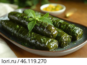 A dish consisting of dolma - grape leaves stuffed with a filling, probably rice, herbs and spices. The leaves look smooth and shiny, covered with fresh herbs and topped with a sprig of mint. The dolma is placed on a dark ceramic plate, creating a contrast with the bright green color of the leaves.A bowl of lemon wedges is slightly blurred in the background, hinting at the traditional accompaniment to the dish - serving with lemon juice for a tart taste. The background is decorated in warm wooden tones, creating a cozy atmosphere of Eastern or Mediterranean cuisine. Стоковое AI-изображение, AI-художник Анна Кривицкая / Фотобанк Лори