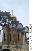 An Orthodox church under construction with a golden dome and an Orthodox cross on top. The walls of the building are made of red brick, in some places not yet covered with a finishing layer. The architectural style is reminiscent of traditional Russian churches, with characteristic semicircular arches above the windows and entrance. Стоковое фото, фотограф Анна Кривицкая / Фотобанк Лори