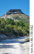 A picturesque Crimean landscape with a dirt road leading to a massive flat-topped rock. This mountain features steep cliffs and layered limestone rocks, which is typical of many Crimean mountain ranges. The lower part of the mountain is covered with dense green trees and bushes, creating a contrast with the dry, rocky slope.Mountain definition:The image probably depicts Mount Kyz-Kermen, one of the notable Crimean flat-topped mountains located near Bakhchisaray. This is an ancient cave settlement, used in the Middle Ages as a fortified settlement. The mountain has expressive rock ledges and grottoes, which can be seen in the photograph. Стоковое фото, фотограф Анна Кривицкая / Фотобанк Лори