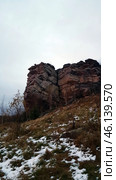 A rock with a characteristic layered structure surrounded by autumn nature. In the foreground, bushes with yellowed leaves are visible, as well as islands of snow on the ground, indicating late autumn or early winter. The rock has a rough texture, many cracks and stratifications, which makes it a unique geological formation.The landscape is typical for the Perm region, where the Ural mountain ranges are combined with forests and open landscapes. Possible location - Vetlan Rock, Chertovo Gorodishche or Stone City. Стоковое фото, фотограф Анна Кривицкая / Фотобанк Лори