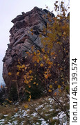 A rock with a characteristic layered structure surrounded by autumn nature. In the foreground, bushes with yellowed leaves are visible, as well as islands of snow on the ground, indicating late autumn or early winter. The rock has a rough texture, many cracks and stratifications, which makes it a unique geological formation.The landscape is typical for the Perm region, where the Ural mountain ranges are combined with forests and open landscapes. Possible location - Vetlan Rock, Chertovo Gorodishche or Stone City. Стоковое фото, фотограф Анна Кривицкая / Фотобанк Лори