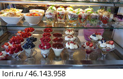 A display case with various desserts and salads arranged in two rows. In the bottom row are bowls with various desserts: with strawberries, blueberries, raspberries, whipped cream, panna cotta and berry sauces. In the top row are portioned salads in glass goblets and white plates, decorated with greens, sliced ​​vegetables, meat, cheese and sauces. In the center of the frame, the multi-layered salads with sliced ​​cucumbers, tomatoes, greens and ham stand out. The whole composition looks appetizing, colorful and elegant. Стоковое фото, фотограф Анна Кривицкая / Фотобанк Лори