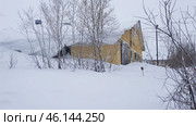 Snow-covered landscape of Vorkuta with a partially snow-covered building. Judging by its appearance, it is an abandoned or rarely used building, possibly an old warehouse, hangar or garage. The snow has formed thick drifts that reach almost to the roof. The branches of the bushes are covered with frost, creating a feeling of a deep winter cold.The building is painted yellowish-brown, contrasting with the whiteness of the snow and the gray sky. Lighting poles and power lines are visible in the distance, indicating an industrial or economic zone of the city.This photo illustrates the harsh conditions of the Far North, where blizzards and snowfalls can literally bury buildings under snowdrifts. Стоковое фото, фотограф Анна Кривицкая / Фотобанк Лори