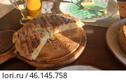 Traditional dish - khachapuri, served on a wooden board. Khachapuri is a Georgian flatbread filled with melted cheese and sometimes egg. You can see the crispy, browned crust with a characteristic pattern from the grill or oven, and inside is stretchy, melted cheese. In the background you can see a plate with leftovers, a glass of water and a glass of orange juice. The atmosphere of the photo conveys coziness and warm, sunny light. Стоковое фото, фотограф Анна Кривицкая / Фотобанк Лори