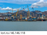 Novorossiysk Commercial Sea Port, one of the largest ports in Russia, located on the Black Sea coast. The frame shows cargo ships, port cranes, container yards and part of the industrial infrastructure. In the background are the mountain ranges surrounding the city.Numerous blue and yellow gantry cranes used for loading and unloading ships.Cargo containers stored in the port area.Commercial ships at the berths undergoing cargo operations.Tugboats used to maneuver large ships.Mountains, characteristic of the Novorossiysk landscape.Bright blue sky with clouds, creating a contrast with the industrial landscape. (2023 год). Редакционное фото, фотограф Анна Кривицкая / Фотобанк Лори