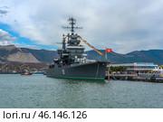 The cruiser Mikhail Kutuzov, permanently moored at the Novorossiysk embankment. This is one of the main museum ships of Russia, which is part of the Central Naval Museum.The cruiser Mikhail Kutuzov is a massive warship with the number "105" on board.It is decorated with signal flags, creating a festive atmosphere.In the background, the port of Novorossiysk, the mountains of the Markotkhsky Range and the berthing facilities are visible.The red naval flag with a white star flutters at the stern.To the right of the ship is the marine terminal, where people and city infrastructure are visible.In the distance, a small tug or boat moves along the water. (2023 год). Редакционное фото, фотограф Анна Кривицкая / Фотобанк Лори
