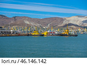 The port of Novorossiysk with its characteristic industrial facilities and infrastructure. In the center of the frame are yellow tugboats moored at the berth. In the background are cargo cranes, container platforms, and the mountains of the Markotkhsky Range with limestone quarries.The port of Novorossiysk is the largest sea trade port in Russia.The yellow tugboats are auxiliary vessels for maneuvering large ships.Cargo cranes - handle containers and cargo.Container terminal - stores sea freight.The mountains of the Markotkhsky Range - form the background of the photo.Bright blue sky with thin cloud stripes.Turquoise water of the Black Sea with light waves. (2023 год). Редакционное фото, фотограф Анна Кривицкая / Фотобанк Лори