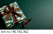 The concept of Defender of the Fatherland Day on February 23. Gift camouflage box with a festive satin ribbon bow on a dark green background. Стоковое AI-изображение, AI-художник Photojuli / Фотобанк Лори