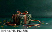 The concept of Defender of the Fatherland Day on February 23. Gift camouflage box with a festive satin ribbon bow on a dark green background. Стоковое AI-изображение, AI-художник Photojuli / Фотобанк Лори
