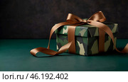 The concept of Defender of the Fatherland Day on February 23. Gift camouflage box with a festive satin ribbon bow on a dark green background. Стоковое AI-изображение, AI-художник Photojuli / Фотобанк Лори