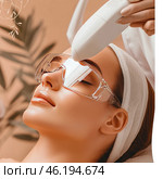 A woman undergoing a cosmetology procedure using a laser or photoepilation device. She is relaxed, her hair is tucked under a white bandage, and her eyes are protected by special transparent glasses. The cosmetologist uses a professional device aimed at the forehead or eyebrow area. The background is decorated in warm natural tones, creating a feeling of coziness and relaxation. Стоковое AI-изображение, AI-художник Анна Кривицкая / Фотобанк Лори