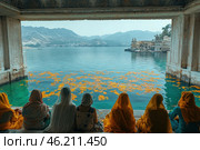 A group of women dressed in traditional saris sit by the water in a carved stone pavilion, watching over the lake. Their clothes are painted in soft pastel and gold tones, in harmony with the surrounding landscape. Bright orange flowers float on the surface of the water, creating a sense of ritual or ceremony. In the background, a majestic palace or temple is visible, located on the shore of the lake, and beyond it, mountain ranges shrouded in mist. The atmosphere of the scene is filled with peace, contemplation and a deep cultural connection with nature and tradition. Стоковое AI-изображение, AI-художник Анна Кривицкая / Фотобанк Лори