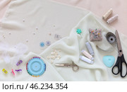 Flat lay of sewing accessories and materials for sew and cutting on white silk fabric - threads, needles, buttons, scissors, tailor chalk, pincushion, needle threader. Sewing as a hobby. Top view, close-up. Стоковое фото, фотограф Виктория Катьянова / Фотобанк Лори
