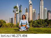 A woman sits cross-legged on green grass, practicing mindfulness against a backdrop of towering skyscrapers. The warm sun bathes the urban landscape in golden light as she finds her peace. Стоковое фото, фотограф Aleksandr Golubev / Фотобанк Лори