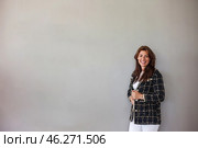A confident woman with long, flowing hair stands beside a smooth gray wall, dressed in an elegant blazer and white pants. Her cheerful smile adds warmth to the minimalist atmosphere of the room. Стоковое фото, фотограф Aleksandr Golubev / Фотобанк Лори