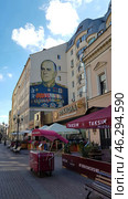 One of the streets of Moscow with a pedestrian area decorated with benches, flowers and street cafes. On the facade of the building there is a large wall graffiti or mural depicting a famous Soviet military leader in a ceremonial uniform with numerous orders and medals. Under the mural is the cafe "Rukkola" with a sign in Russian and the neighboring restaurant "Taksim", specializing in Turkish cuisine. In the foreground is a mobile stall with a pink awning. The background includes light historical buildings typical of the center of Moscow, as well as a blue sky with clouds. (2018 год). Редакционное фото, фотограф Анна Кривицкая / Фотобанк Лори