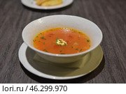 A plate of tomato soup, perhaps borscht or tomato soup with vegetables. In the center of the soup is a small piece of sour cream or butter, which gradually dissolves. The soup is garnished with herbs, perhaps parsley or dill. The plate is on a white saucer, with another plate with bread or croutons visible in the background. Стоковое фото, фотограф Анна Кривицкая / Фотобанк Лори
