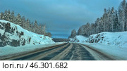 A winter road running through a snow-covered forest. On both sides of the road are snow banks and exposed rock formations covered in a thick layer of snow. The road is partially cleared of snow, but is covered with ice and mud from cars, which gives it a characteristic winter look. In the distance, the road smoothly disappears into the horizon, surrounded by tall snow-covered trees. The sky is painted in cold blue and gray shades, creating an atmosphere of winter travel and tranquility. Стоковое фото, фотограф Анна Кривицкая / Фотобанк Лори