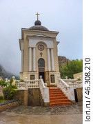 Chapel of the Nativity of Christ, located in the Laspi tract, Crimea. This is a small Orthodox chapel with classical architecture: a light facade, four columns at the entrance, a semicircular dome with a cross on top. Above the entrance is an image of Jesus Christ, and below it is a golden cross. A staircase with terracotta steps, framed by white railings with decorative balls, leads to the entrance. The open door of the chapel symbolically invites to prayer and spiritual reflection. (2021 год). Редакционное фото, фотограф Анна Кривицкая / Фотобанк Лори