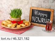 Chalkboard with text HAPPY NOWRUZ, treats, grass and glass of tea on table near grunge wall. Стоковое фото, фотограф Pixel_shot / Фотобанк Лори