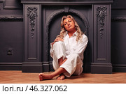 A 45-year-old woman sits comfortably on the floor, dressed in casual white attire, immersed in a serene moment amidst a tasteful indoor setting with rich, dark hues. Стоковое фото, фотограф Aleksandr Golubev / Фотобанк Лори