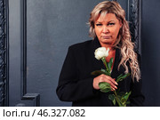 A middle-aged woman, exuding confidence, stands indoors against a textured wall, holding a white rose. The atmosphere is intimate, reflecting both elegance and strength in her demeanor. Стоковое фото, фотограф Aleksandr Golubev / Фотобанк Лори