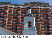 Contrast between the historical and modern architecture of Moscow. In the foreground is the bell tower of an ancient church, painted in blue and white, topped with an onion-shaped dome with a cross. Church bells hang in the opening of the tower.In the background rises the President Hotel, a monumental Soviet building with its characteristic beige brick cladding, many windows and cascading structure.This image perfectly demonstrates the coexistence of religious heritage and Soviet modernism, typical of Moscow. Стоковое фото, фотограф Анна Кривицкая / Фотобанк Лори
