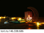 Arabic candle holder with glowing candle, garland, Quran and tasbih on table against dark background. Стоковое фото, фотограф Pixel_shot / Фотобанк Лори