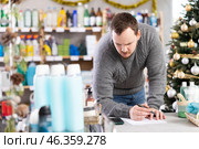 Attentive male salesperson restock of goods, checking papers in small household store decorated for New Year celebration. Стоковое фото, фотограф Яков Филимонов / Фотобанк Лори