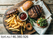 French fries, burger, pork ribs on a stylish board on a wooden background. Fried potatoes. The concept of fast food, junk food and fast food preparation. Стоковое AI-изображение, AI-художник Photojuli / Фотобанк Лори